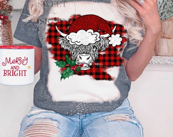 May include: A gray t-shirt with a distressed design featuring a Highland cow wearing a Santa hat and holding a sprig of holly. The background is a red and black buffalo plaid pattern. The text "Merry and Bright" is on a red mug.