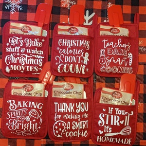 May include: Set of six red oven mitts with white text and graphics. The mitts feature various Christmas-themed sayings, including "Let's Bake Stuff & Watch Christmas Movies", "Christmas Calories Don't Count", "Teachers Bake Smart Cookies", "Baking Spirits Bright", "Thank You for Making Me a Smart Cookie", and "If You Stir It, It's Homemade".