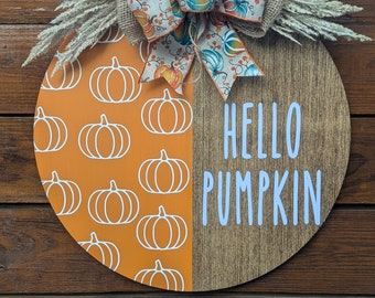 May include: A round wooden fall-themed sign with a burlap bow and wheat stalks. The left side is orange with white pumpkin outlines, and the right side is wood-grain with the words "HELLO PUMPKIN" in white.