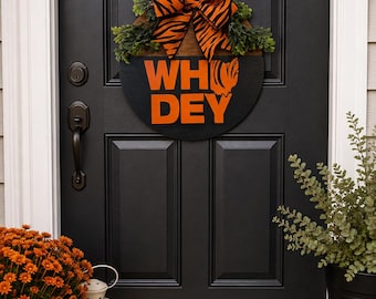 May include: A circular wooden door sign featuring the orange text WHO DEY with a tiger stripe pattern on the letters. The sign is topped with a large orange and black tiger-striped fabric bow and green faux foliage accents, hanging by a rope.
