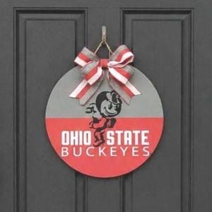 May include: A circular wooden door sign featuring a horizontal split design with a grey top half and a red bottom half. The text OHIO STATE BUCKEYES is printed in white, accompanied by a black mascot graphic. A striped red, white, and grey bow is attached.