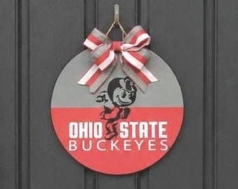 May include: A circular wooden door sign featuring a horizontal split design with a grey top half and a red bottom half. The text OHIO STATE BUCKEYES is printed in white, accompanied by a black mascot graphic. A striped red, white, and grey bow is attached.