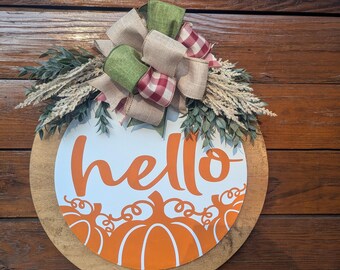 May include: A round wooden sign with the word "hello" in orange script. The sign features orange pumpkin graphics and is decorated with a bow made of green, tan, and red plaid ribbons, along with wheat and greenery. The sign is a fall-themed decoration.