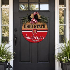 May include: A round wooden door sign featuring the text OHIO STATE in black block letters and buckeyes in white script. The sign is divided into a wood-toned top and a red bottom with a central O logo, topped with a plaid and burlap bow.