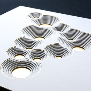 Op de afbeelding: Witte papierkunst met een gouden omtrek, met een ontwerp van overlappende cirkels met concentrische ringen. De cirkels zijn uit het papier gesneden, waardoor een 3D-effect ontstaat.