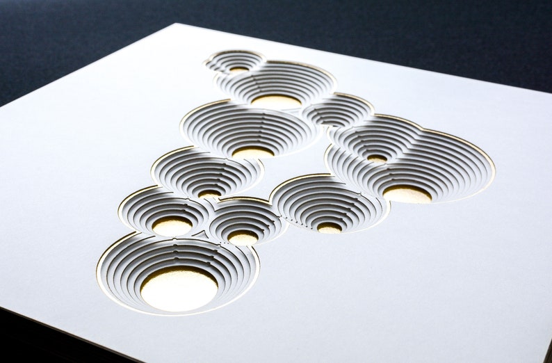 Op de afbeelding: Witte papierkunst met een gouden omtrek, met een ontwerp van overlappende cirkels met concentrische ringen. De cirkels zijn uit het papier gesneden, waardoor een 3D-effect ontstaat.