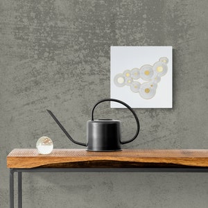Op de afbeelding: Een zwarte gieter met een lange tuit staat op een houten tafel met een heldere glazen bol. Een wit vierkant canvas met een gouden en wit abstract ontwerp hangt aan een grijze, getextureerde muur achter de tafel.