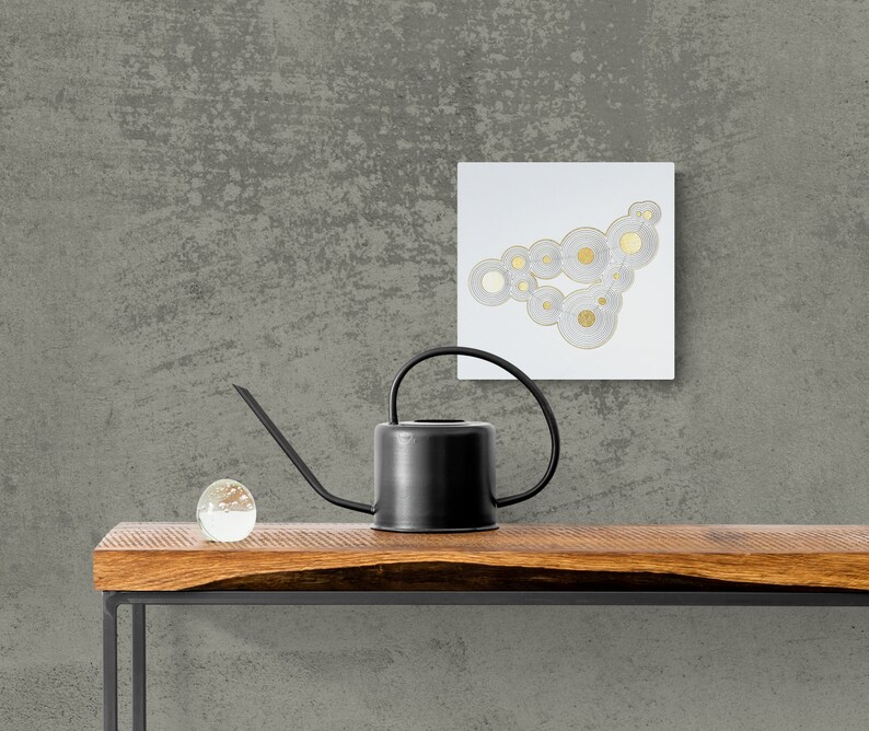 Op de afbeelding: Een zwarte gieter met een lange tuit staat op een houten tafel met een heldere glazen bol. Een wit vierkant canvas met een gouden en wit abstract ontwerp hangt aan een grijze, getextureerde muur achter de tafel.