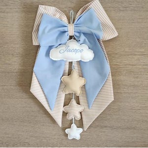 Può includere: Un fiocco di stoffa a righe blu chiaro e beige con una nuvola bianca che dice "Jacopo" appesa ad esso. Il fiocco ha una catena di stelle appese ad esso, tra cui una stella beige, una stella marrone e una stella bianca.