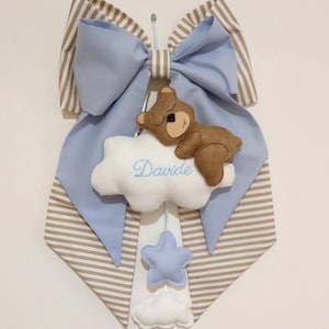 Può includere: Decorazione per la cameretta dei bambini con un grande fiocco blu e dettagli a righe marroni e bianche. Un orsacchiotto marrone riposa su una nuvola bianca con il nome "Davide" in corsivo blu. Include stelle e nuvole blu.