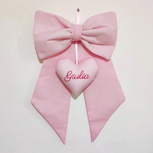Può includere: Un fiocco di stoffa rosa con un cuscino a forma di cuore appeso. Il cuscino è ricamato con il nome "Giulia".