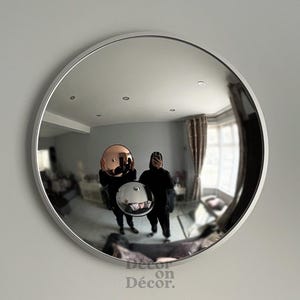 May include: A round, convex mirror with a white frame. The mirror reflects a room with two people holding spherical, reflective objects. The objects are a copper and a silver color. The text "Decor on Décor" is at the bottom.