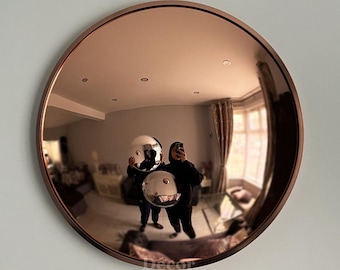 May include: A large, round mirror with a copper-colored frame. The mirror reflects a room with furniture and two people holding reflective spheres. The mirror is mounted on a light gray wall. The text "Décor on Décor" is visible at the bottom.