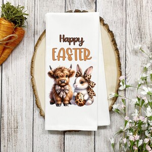 May include: White kitchen towel with a brown Highland cow and a white bunny rabbit holding an Easter egg. The text "Happy Easter" is printed in orange on the towel.