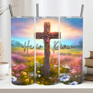 以下が含まれることがあります： カラフルな空の下、ピンクの花で飾られた木の十字架が、鮮やかな野草の野原を背景にした、ラップアラウンドデザインのステンレススチールタンブラー。「He is Risen」とマタイ28:6のテキストも表示されています。