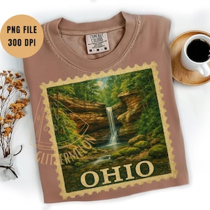 May include: A dusty rose Comfort Colors t-shirt featuring a scenic Ohio waterfall design within a postage stamp border. The shirt includes the text "OHIO" in a gold color. A coffee cup and saucer are on the right.