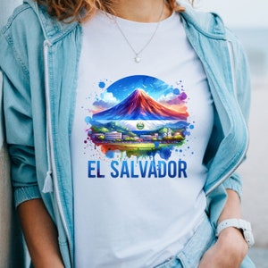 May include: White t-shirt featuring a colorful illustration of El Salvador. The design includes a mountain, the Salvadoran flag, and the text "EL SALVADOR" in blue. The shirt is worn with a light blue jacket.