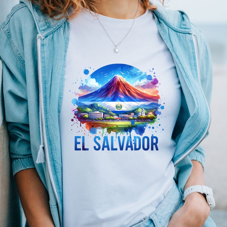 May include: White t-shirt featuring a colorful illustration of El Salvador. The design includes a mountain, the Salvadoran flag, and the text "EL SALVADOR" in blue. The shirt is worn with a light blue jacket.