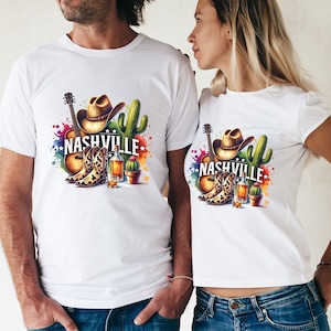Pu&ograve; includere: Maglietta bianca con un design grafico colorato che presenta un cappello da cowboy, stivali, una chitarra e un cactus. Il testo "NASHVILLE" &egrave; stampato in lettere nere.