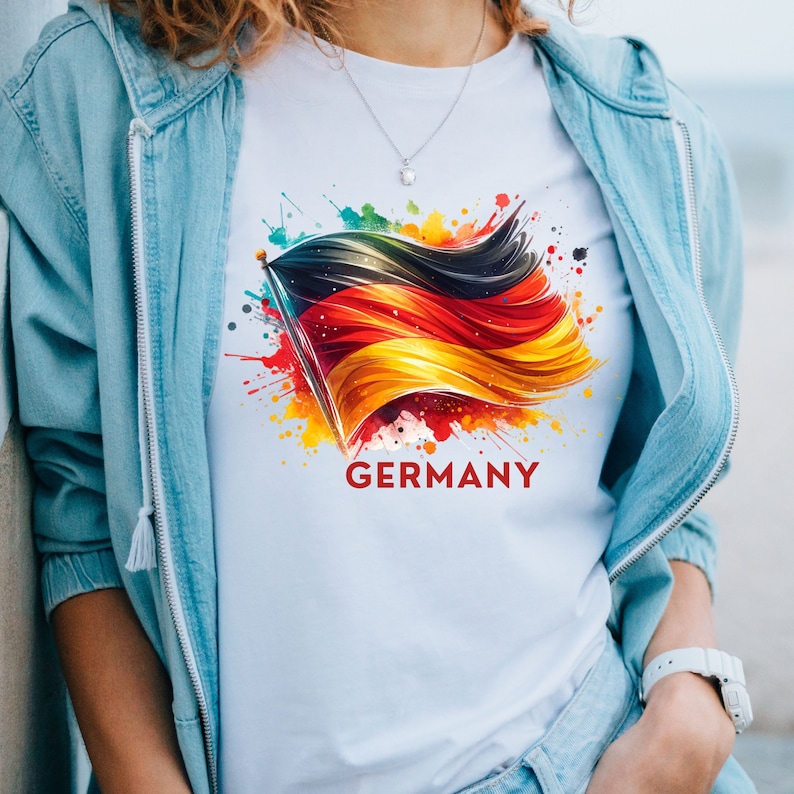 Puede incluir: Camiseta blanca con un dise&ntilde;o de acuarela colorido de una bandera alemana ondeando al viento. El texto "GERMANY" est&aacute; impreso debajo de la bandera.