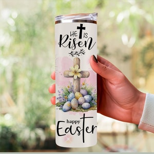 May include: A white stainless steel tumbler with a lid, featuring the text "He is Risen" and "Happy Easter". The design includes a wooden cross, Easter eggs, and floral accents in pastel colors.