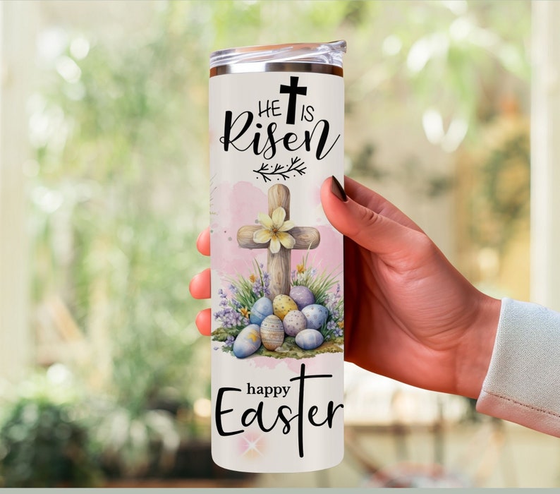 May include: A white stainless steel tumbler with a lid, featuring the text "He is Risen" and "Happy Easter". The design includes a wooden cross, Easter eggs, and floral accents in pastel colors.