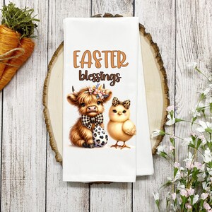 May include: White kitchen towel with a brown Highland cow and a yellow chick. The cow is wearing a black and white bow tie and a floral headband. The chick is wearing a black and white bow. The text "Easter Blessings" is printed in brown above the animals.