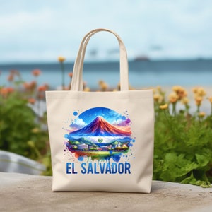 May include: A cream-colored tote bag featuring a watercolor design of a mountain, the El Salvador flag, and the text "EL SALVADOR" in blue. The bag has two cream-colored handles and is sitting on a stone surface.