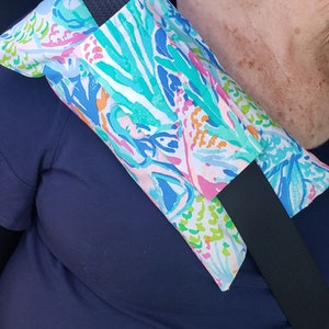 May include: A colorful patterned seatbelt shoulder pad with a black seatbelt strap. The pad is made of fabric and has a floral design with pink, blue, green, and white colors.