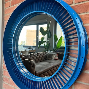 May include: A round, blue mirror with a unique industrial design. The mirror's frame features a series of angled blades, creating a turbine-like appearance. The mirror is mounted on a red brick wall, reflecting a room with a brown sofa and large windows.