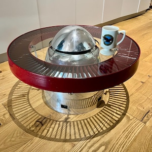 Könnte beinhalten: Ein einzigartiger Couchtisch mit rotem Rand und Glasplatte. Der Tisch hat eine zentrale, metallische, turbinenartige Struktur. Ein weißer Becher mit blau-grüner Grafik steht auf der Glasoberfläche.