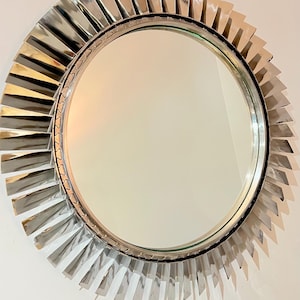 May include: A round mirror with a silver frame that resembles a jet engine. The frame is made of many thin, silver blades that radiate outwards from the center of the mirror.