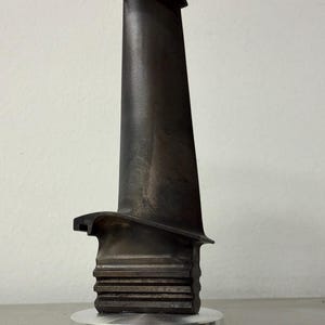Könnte beinhalten: Eine dunkelgraue, metallische Turbinenschaufel mit einem glatten, länglichen Profil und einem gerippten, rechteckigen Fuß. Das Bauteil ist vertikal auf einem runden, gebürsteten silbernen Metallsockel montiert und zeigt industrielles Design.