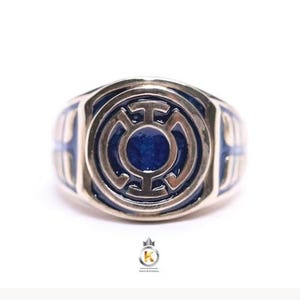 May include: A metallic ring featuring a circular face with a raised, stylized emblem set against a deep blue enamel background. The band is polished metal with matching blue enamel accents along the sides, creating a cohesive and detailed design.