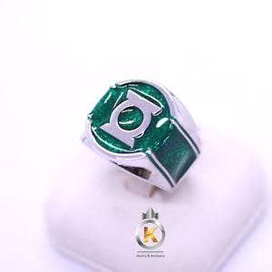 May include: A silver-toned ring featuring a circular face with a green, textured inlay and a prominent silver Green Lantern logo symbol centered on the top. The band has matching green textured side panels and a polished metallic finish throughout.