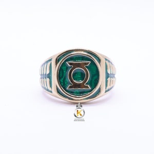 May include: Gold-coloured ring with a green enamel emblem featuring a stylized design. The ring has a rounded shape with a detailed band. The emblem is a prominent focal point, set against a white background. The ring is a piece of jewellery.