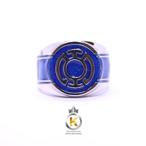 Può includere: Un anello color argento con una faccia circolare e un inserto in smalto blu metallizzato. Al centro dell'anello è presente un simbolo argentato in rilievo che ricorda l'icona di una lanterna stilizzata. La fascia è liscia e lucida.