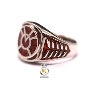 May include: A silver ring with a red enamel design. The design features a circle with a stylized symbol inside. The ring has a textured band.