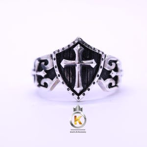 Könnte beinhalten: Ein silberfarbener Ring mit einem zentralen schildförmigen Emblem und einem erhabenen Kreuzdesign. Der Schild hat einen dunklen, vertieften Hintergrund und einen gepunkteten Rand. Die Ringschiene zeigt auf beiden Seiten komplexe Schnörkelmuster.