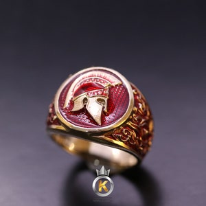 May include: A gold ring with a red enamel design featuring a Spartan helmet. The ring has intricate details and a gold band.