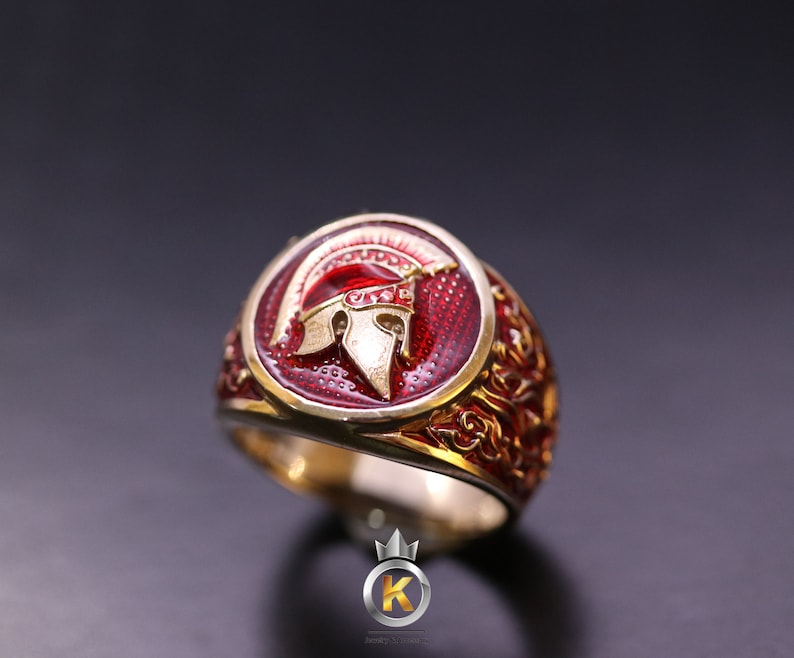 May include: A gold ring with a red enamel design featuring a Spartan helmet. The ring has intricate details and a gold band.