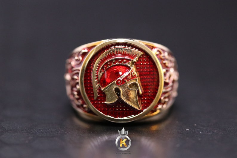 May include: A gold ring with a red enamel design featuring a Spartan helmet. The helmet is gold with a red crest and is set against a red background.