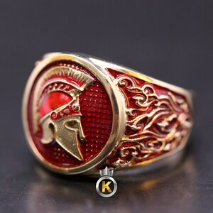May include: A gold ring with a red enamel design featuring a Spartan helmet. The ring has a detailed design with gold accents.