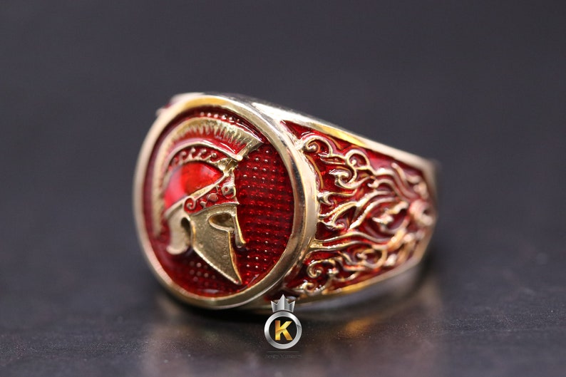 May include: A gold ring with a red enamel design featuring a Spartan helmet. The ring has a detailed design with gold accents.