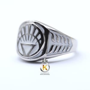 May include: A silver ring with a white enamel design of a stylized eye with a triangle in the center. The ring band has a textured design.