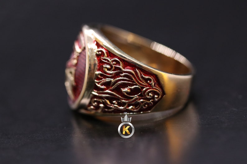 May include: A gold ring with a red enamel design. The ring features intricate details and a raised crest.