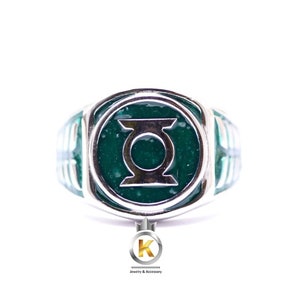 May include: A silver-toned ring featuring a circular face with a dark green, textured inlay and a prominent silver metallic Green Lantern symbol centred on the face. The band has a segmented design with green accents along the sides of the ring.