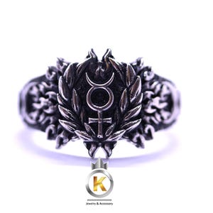 Könnte beinhalten: Silberfarbener Ring mit einem zentralen Emblem mit Mond und Kreuz, umgeben von einem Lorbeerkranz. Der Ring hat aufwendige Details und ein strukturiertes Band. Ein Schmuckaccessoire.