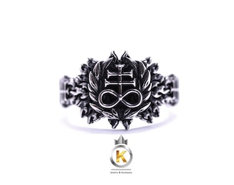May include: A silver-toned ring featuring a central emblem with an inverted cross and an infinity symbol, surrounded by a detailed floral or leaf design. The ring has a wide band with intricate patterns.