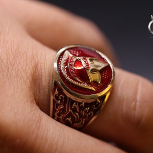 May include: A gold ring with a red enamel design featuring a stylized warrior helmet. The ring has intricate details and a polished finish.